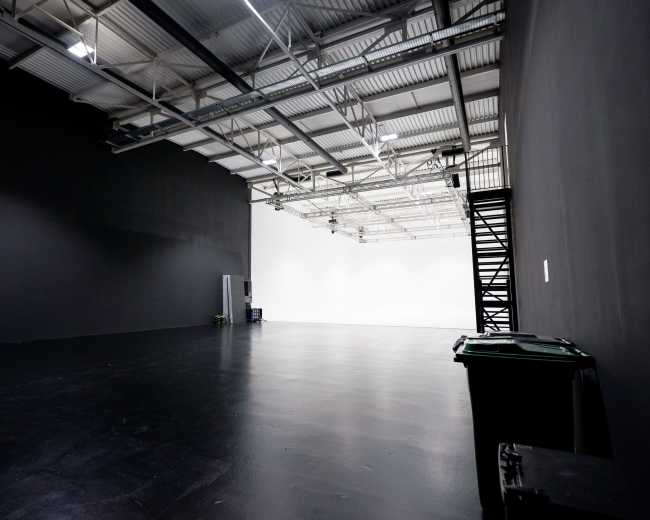 The image shows a spacious studio with a black wall on one side and a white wall on the opposite side, featuring a polished floor and minimal furnishings.