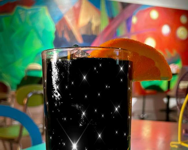 A glass filled with a dark beverage, garnished with an orange slice, is placed on a colorful table.