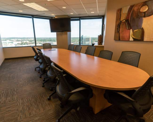 The image shows a modern conference room featuring a long wooden table surrounded by black office chairs, with large windows providing an expansive view.