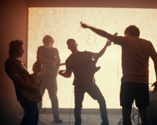 A band performs on stage in front of a bright backdrop, with members playing instruments and singing.