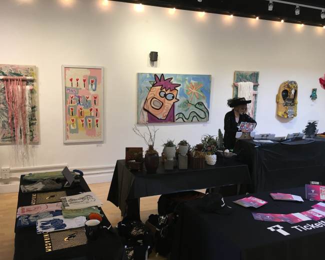 The scene shows an art exhibition with various colorful paintings displayed on the walls and a vendor setting up a table with merchandise.