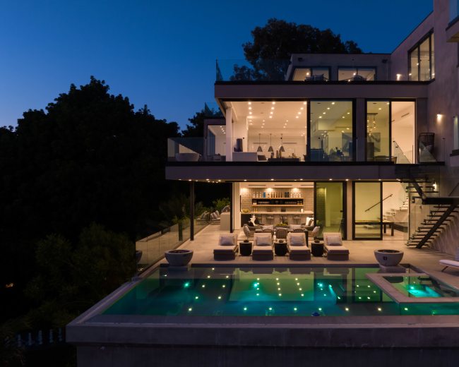 A modern two-story house features large glass windows, an outdoor pool with illuminated water, and a spacious patio area furnished with lounge chairs.