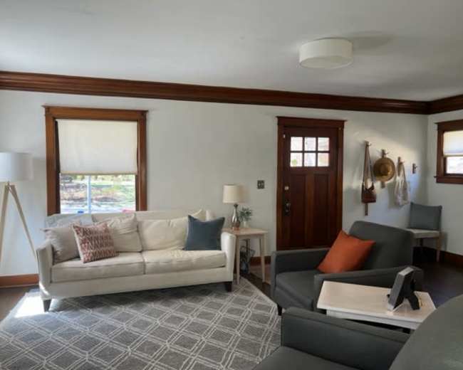 The image shows a living room with a white couch, two armchairs, a lamp, and a front door, all surrounded by light-colored walls and wood trim.
