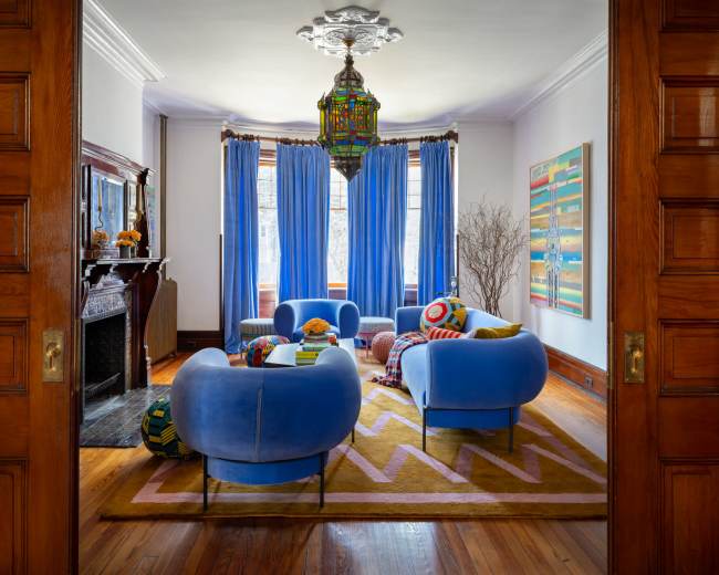 The image shows a well-decorated living room featuring blue sofas, colorful cushions, and large windows with blue curtains.