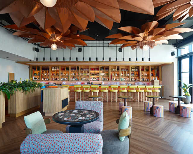A modern bar interior with large floral light fixtures, a wooden bar counter lined with colorful stools, and vibrant plants positioned throughout the space.