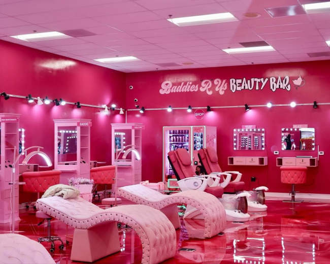 The image shows a brightly lit beauty bar featuring pink decor, plush seating, and vanity stations with mirrors.