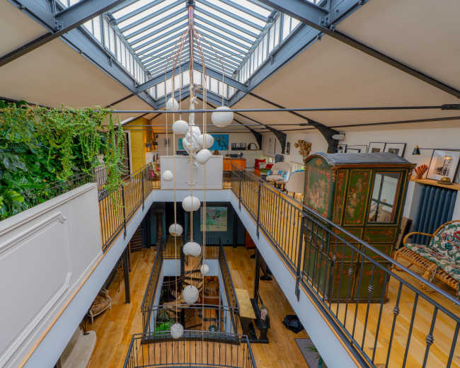The image shows an open, spacious loft interior with a glass ceiling, plants cascading down, and a decorative arrangement of hanging lights.