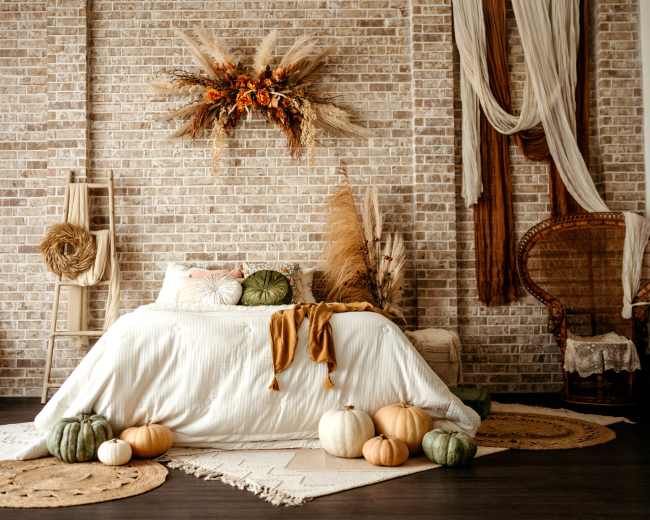 A cozy bedroom features a bed adorned with a white bedspread, surrounded by decorative pumpkins and arranged with natural elements on a textured brick wall.