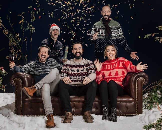 Five people wearing festive sweaters celebrate on a couch with confetti falling around them in a winter-themed setting.