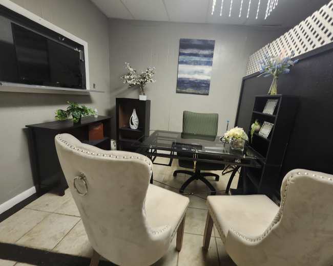 The image shows a small office space featuring a glass-top desk, two upholstered chairs, a decorative wall shelf, and a television mounted on the wall.