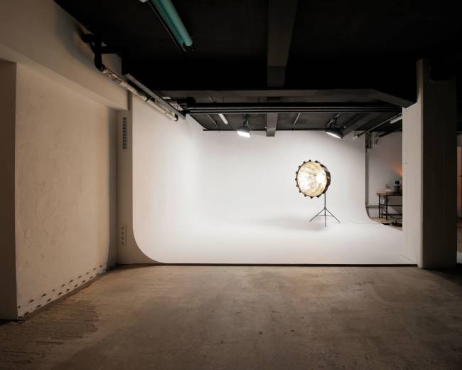 A photography studio features a circular light source on a stand in front of a white backdrop, surrounded by concrete walls and a dark ceiling.