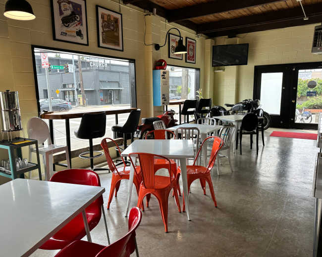 The image shows a modern café interior with a mix of red and white chairs and tables, large windows facing the street, and a motorcycle displayed in the corner.