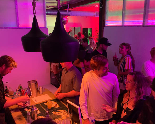 A crowd socializes in a dimly lit room with colorful lighting, while two individuals chat near a bar area.