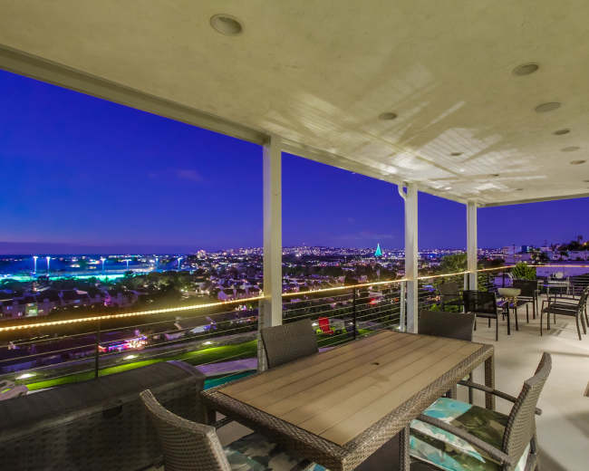 A spacious balcony with a wooden dining table and chairs, overlooking a city skyline illuminated at night.