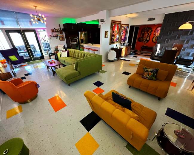 The image shows a vibrant living area with colorful furniture, including green, orange, and yellow sofas, a round coffee table, and artwork on the walls.