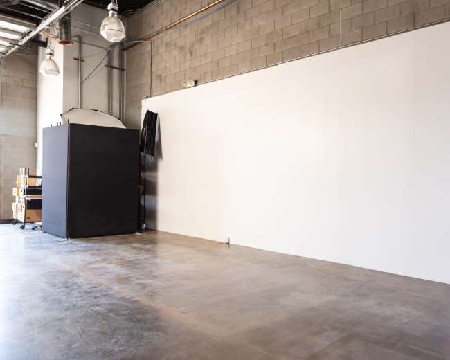 The image depicts a large, empty interior room with a concrete floor and a plain white wall, featuring a dark structure in the corner.