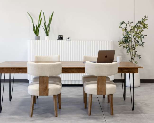 A simple wooden table with four upholstered chairs is set in a minimalistic room featuring a potted plant and a white, ribbed sideboard.