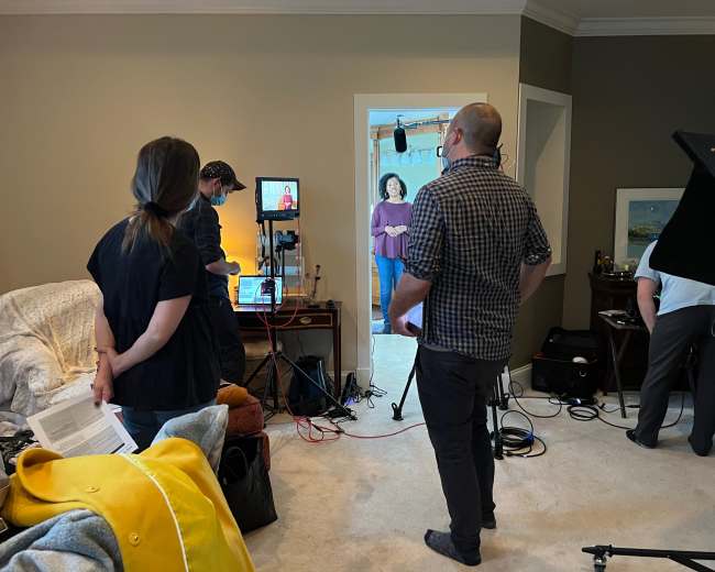 A group of individuals is engaged in a filming setup in a living room, with cameras and equipment arranged in the foreground and a person standing in the doorway.