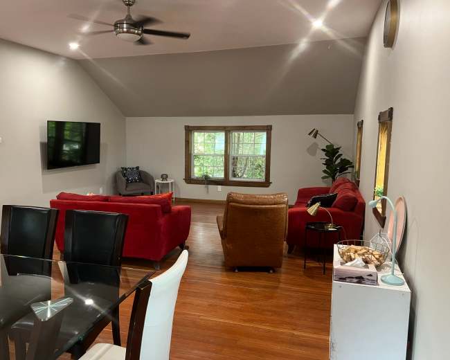 The image shows a living room with a ceiling fan, a large window, red sofas, a brown armchair, and a dining table.