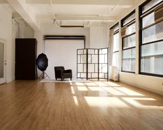 A bright, open studio space features a black chair, a backdrop setup, and large windows casting shadows on the wooden floor.