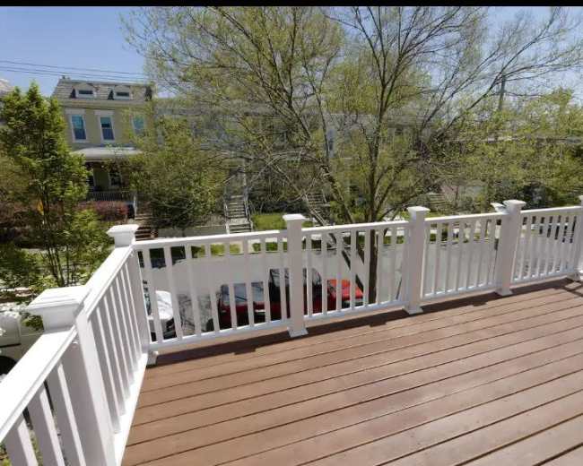 The image shows a wooden deck with white railings overlooking a street lined with parked cars and trees.