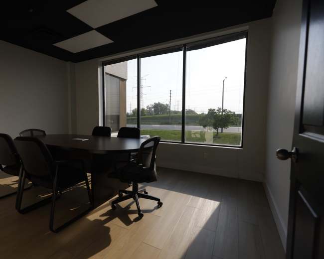 A conference room with a round table and several chairs is illuminated by natural light coming through large windows.