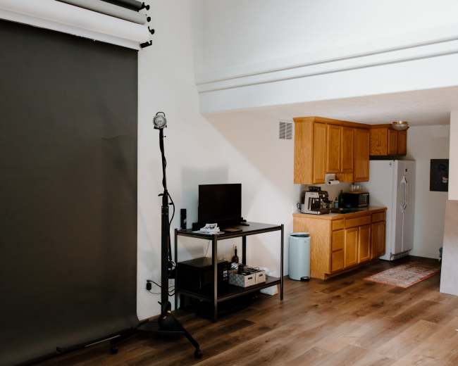 A studio space with a dark backdrop, a television on a cart, and a kitchen area with wooden cabinets and appliances.