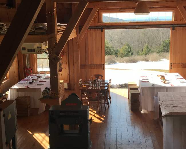 A rustic wooden interior with dining tables set for a gathering, framed by large doors opening to a snowy outdoor landscape.