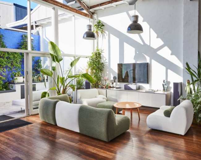 The image shows a modern living room with large windows, featuring green and white furniture, a flat-screen TV, and various plants.