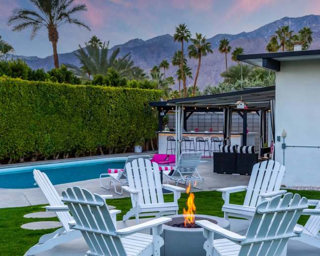 A circular arrangement of white chairs surrounds a fire pit near a pool and a bar area, with mountains in the background and palm trees lining the property.