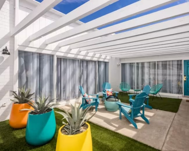A modern outdoor courtyard with colorful seating, potted plants, and a bright blue door under a slatted white roof.