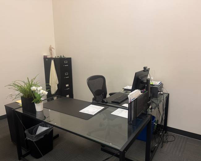 The image shows a minimalist office space featuring a glass desk with a computer, an office chair, a filing cabinet, and a small potted plant.