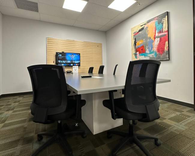 A modern conference room with a large white table, several black office chairs, and a wall-mounted screen, complemented by abstract artwork.