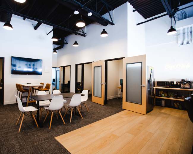 The image shows a modern office space featuring a large table with white chairs, open cubicles, and frosted glass doors.