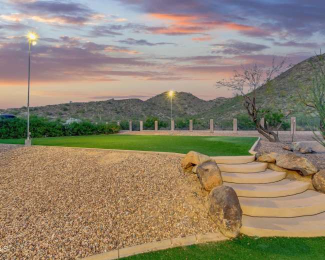 The image shows a landscaped area with stone steps leading up from a gravel surface to a grassy lawn, set against a backdrop of hills and a colorful sunset.