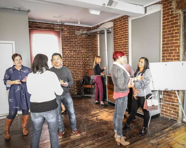 A group of people is gathered in a room with exposed brick walls, engaging in conversation while holding drinks.