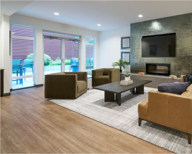The image shows a modern living room with two sofas, a coffee table, and a large television mounted on the wall, alongside large windows that provide natural light.