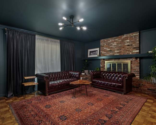 A living room with dark blue walls, a brick fireplace, two deep red leather sofas, a small wooden table, and a large area rug.