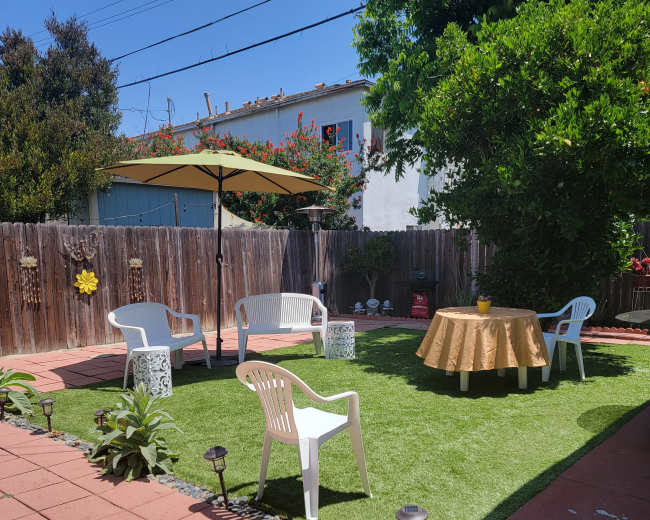 A small backyard features artificial grass, a round table with a gold tablecloth, white chairs, and an umbrella, surrounded by a wooden fence and garden plants.