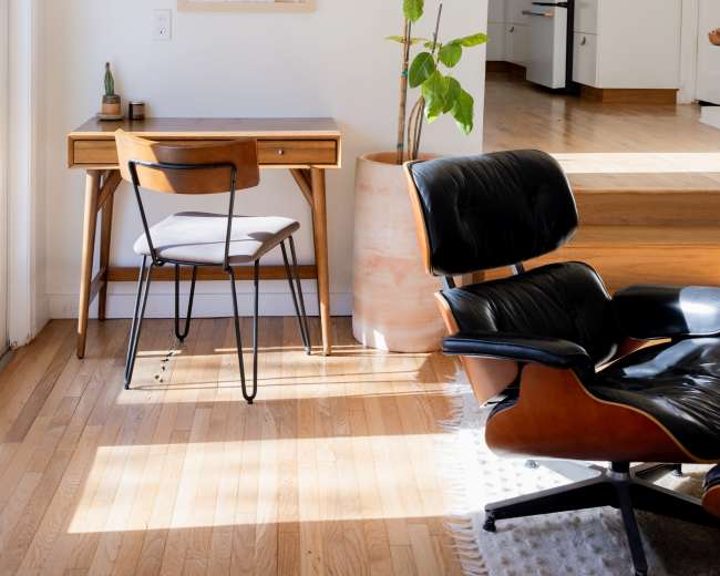 A small desk with a chair is positioned beside a large plant and a map on the wall in a sunlit room leading to a kitchen.