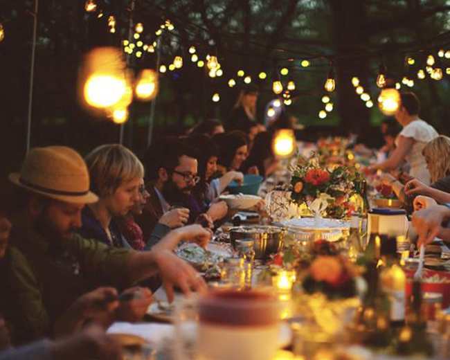 A large group of people sits at a long table adorned with flowers and illuminated by hanging lights during an outdoor dinner.