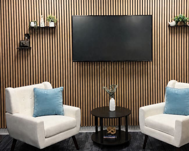 The image shows a modern seating area with two white armchairs featuring blue cushions, positioned around a small circular table, against a backdrop of vertical wooden slats with wall-mounted shelves.