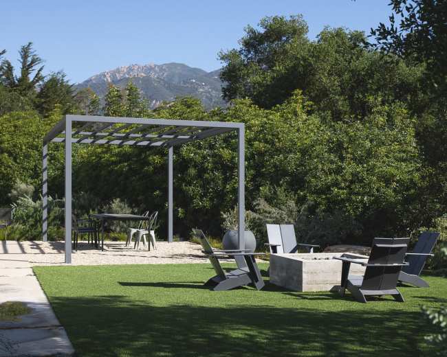 A modern outdoor seating area with a fire pit is set on a grassy lawn surrounded by trees, with a pergola structure in the background and mountains in the distance.