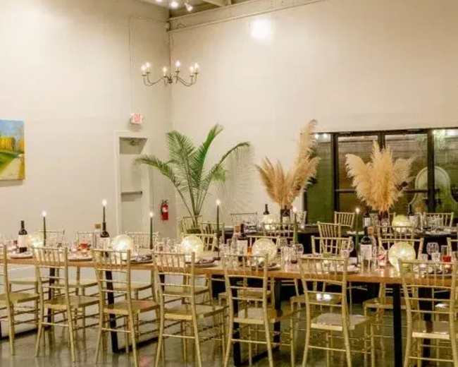 A large dining table is set with candles and decorative plants in a minimalist indoor event space.