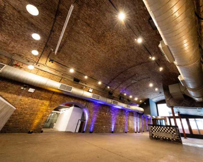 The image shows a spacious industrial-style venue with a brick arch ceiling, exposed ductwork, and ambient lighting along the walls.