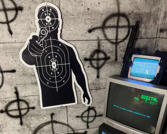 The image shows a corner with a graphic of a shooter on the wall, two old television sets displaying static, and various symbols sprayed as graffiti on a concrete background.