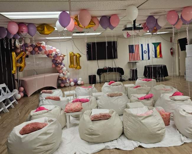 The room features a party setup with bean bags in a circular arrangement, colorful balloons, and flags on the walls.