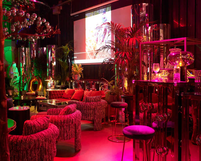 The image shows a vibrant lounge area with colorful lighting, plush seating, and large plants, featuring a screen displaying visuals in the background.