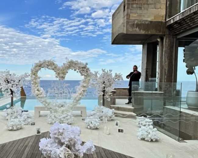 A luxurious outdoor space features a heart-shaped floral arrangement by a pool, with a scenic ocean view in the background.