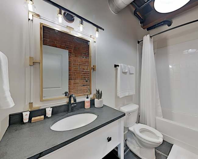 A bathroom featuring a sink with a large mirror, a toilet, a shower with a white curtain, and exposed brick walls.